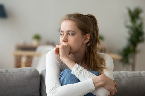 A woman in a white shirt is sitting on a couch looking sideways at a distance. She has her knees up and her arms are wrapped around her knees. She looks anxious. 