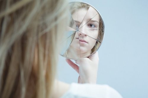Woman examining her reflection in a cracked handheld mirror. Symbolizing the struggle of having bipolar disorder.